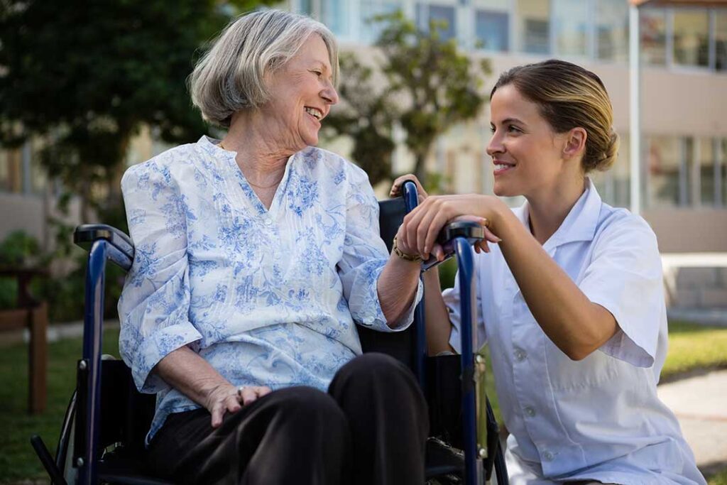 Caregiver helps an older resident walk safely through a bright assisted living hallway.